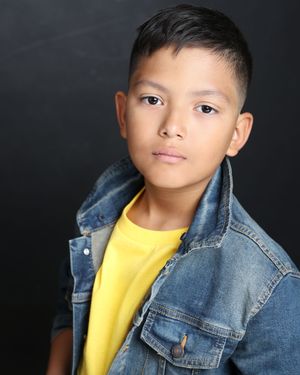 A professional studio portrait with black background showing someone in a yellow shirt wearing a denim jacket.