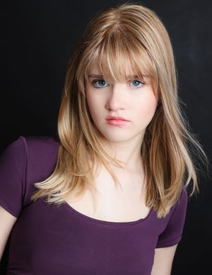 Studio portrait with blonde hair styled with bangs wearing a purple fitted top against dark background.