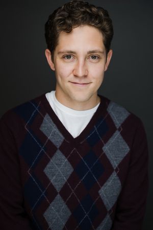Young man smiling in argyle sweater vest over white shirt, professional headshot on dark background.