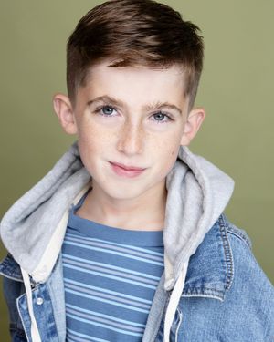 Young boy with blue eyes and freckles wearing a denim jacket and gray hoodie, smiling in a professional headshot.