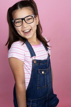 Cheerful young girl wearing glasses and denim overalls smiling against a pink background.