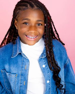 Smiling young girl with box braids and braces wearing a denim jacket over a white turtleneck against a pink background.