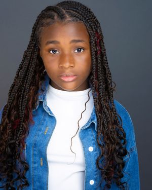 Young girl with long braided hair wearing a denim jacket and white turtleneck poses for a professional headshot.