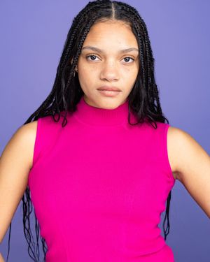 Young woman with long black box braids wearing a bright pink sleeveless turtleneck top against a purple background.