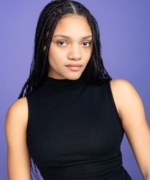Young woman with long box braids wearing a black sleeveless turtleneck top, posing against a purple background.