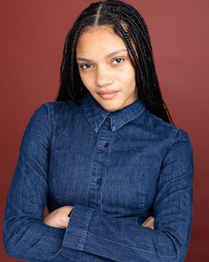 Young woman with long box braids wearing a denim shirt, arms crossed, posing against a dark red background.