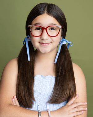 Young girl with red glasses, blue hair ribbons, and pigtails poses with arms crossed against green background.