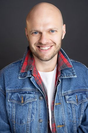 Smiling bald man with blue eyes wearing a denim jacket over a red flannel shirt against a gray background.