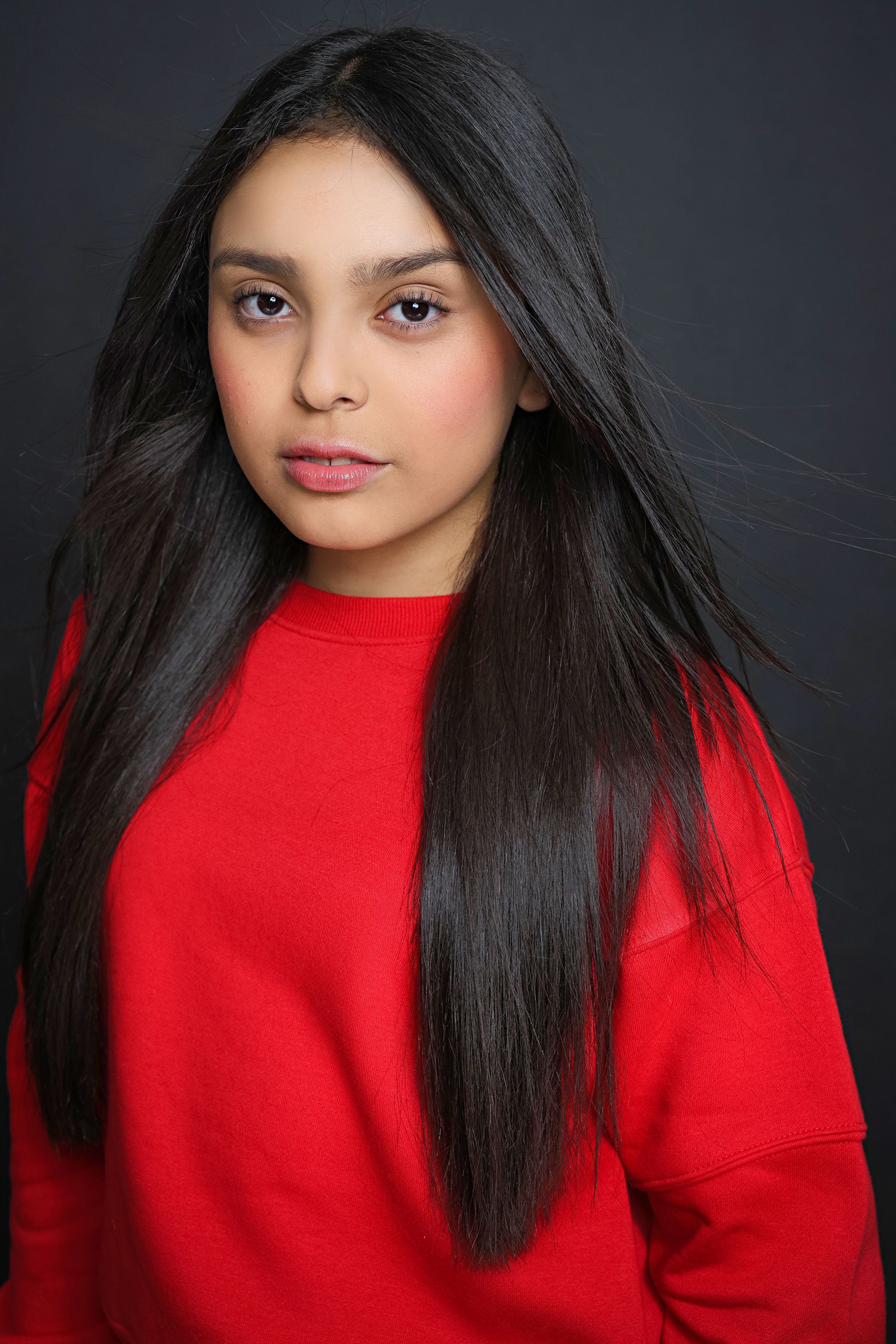 Young girl with long black hair wearing a red sweatshirt poses against a dark gray background.