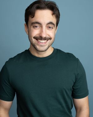 Smiling young man with mustache wearing a dark green t-shirt poses for a professional headshot against a blue background.