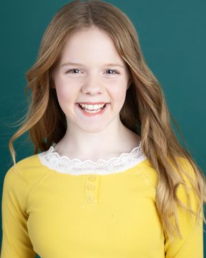 Smiling young girl with long wavy auburn hair wearing a yellow top with white lace trim, posing for a headshot.