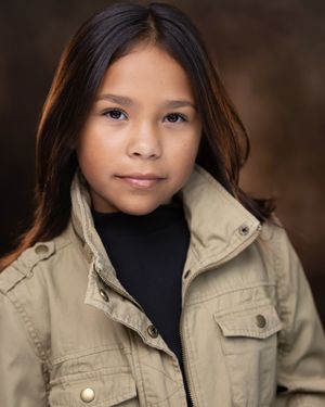 Young girl with long dark hair wearing a tan jacket over a black turtleneck, professional headshot photo.