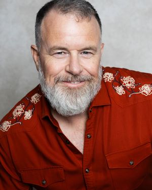 Smiling middle-aged man with gray beard wearing a red western shirt with floral embroidery, professional headshot photo.