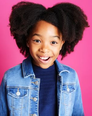 Joyful youth with natural hair styled in puffs wearing a navy sweater and denim jacket against a pink backdrop.