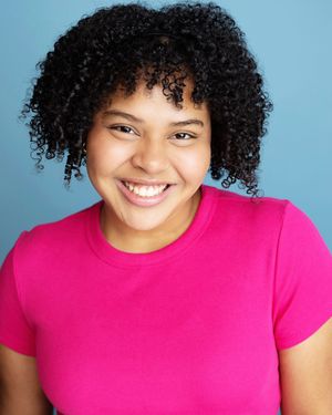 Young woman with curly hair smiling brightly, wearing a pink t-shirt against a blue background.