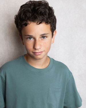 Young boy with curly dark hair wearing a teal t-shirt poses for a headshot photo against a light background.