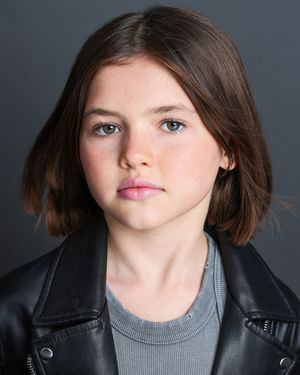 Portrait of a young person with short brown hair wearing a gray top and black leather jacket against a dark background.