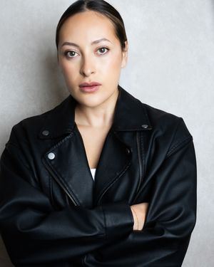 Woman in black leather jacket with arms crossed, serious expression, professional headshot photo.