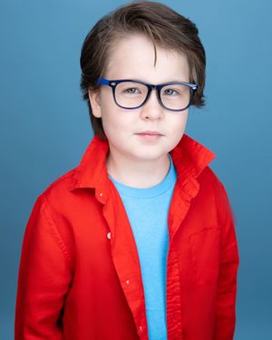 Young boy with blue glasses and red jacket poses for a professional headshot against a blue background.