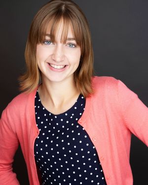 Smiling young woman with brown hair wearing a pink cardigan over a navy polka dot top against a dark background.
