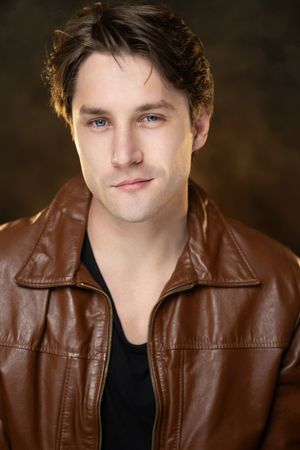 Young man with blue eyes wearing a brown leather jacket and black shirt, posing for a professional headshot photo.
