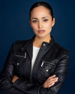 Woman in black leather jacket with arms crossed poses against a dark blue background in a professional headshot.