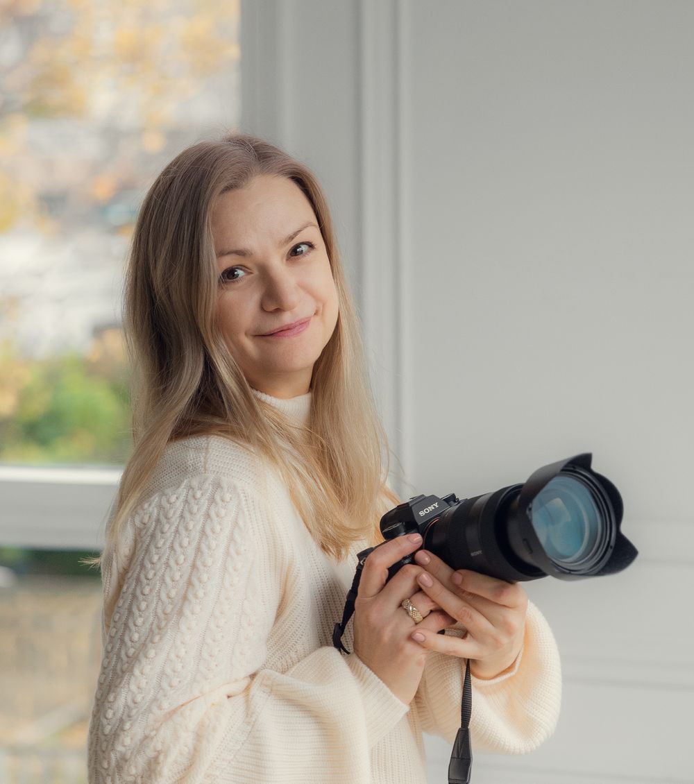 Professional photographer in a white knit sweater holding a DSLR camera by a window.