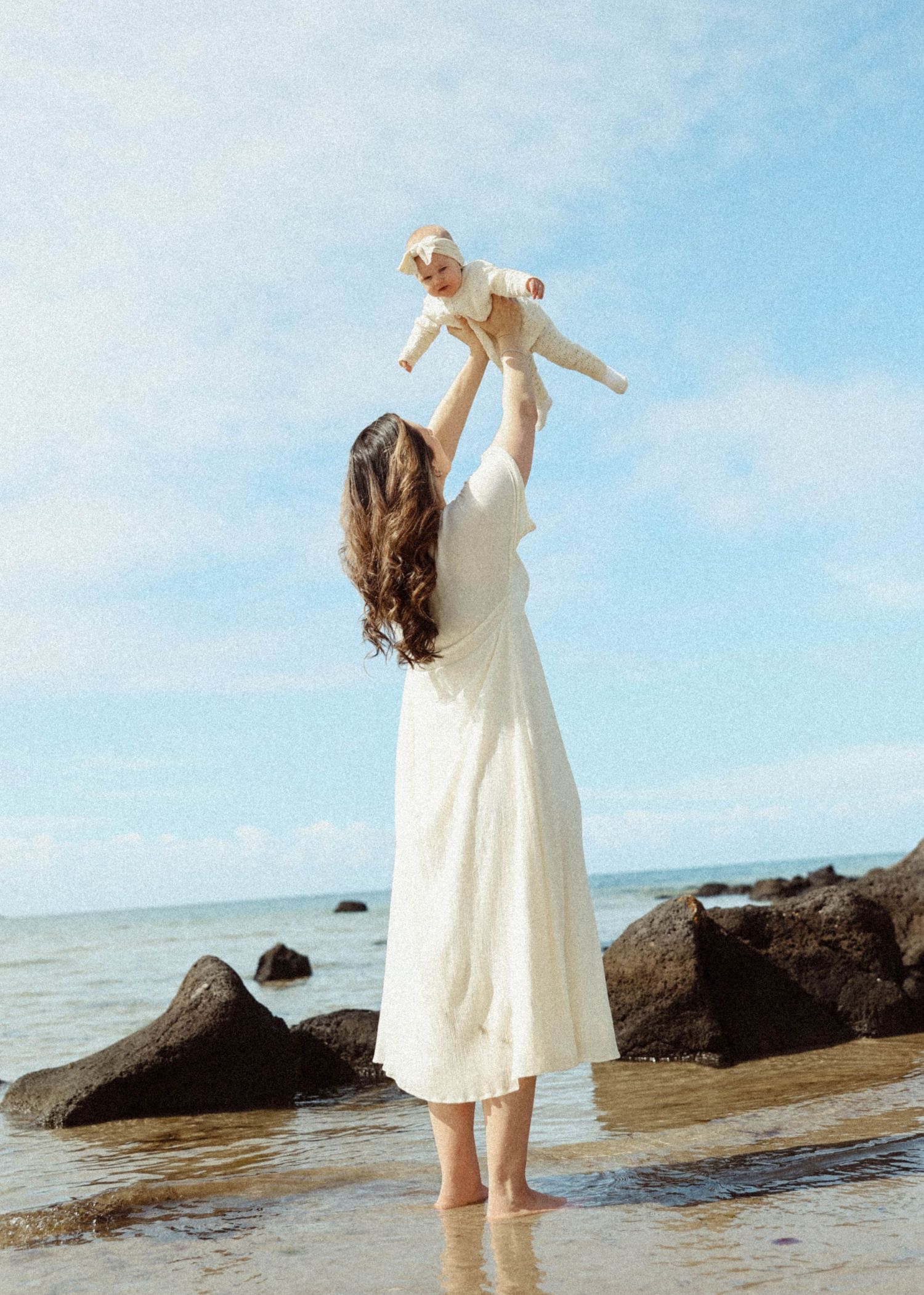 Mother standing barefoot at the beach in a white dress during a natural motherhood photography session.