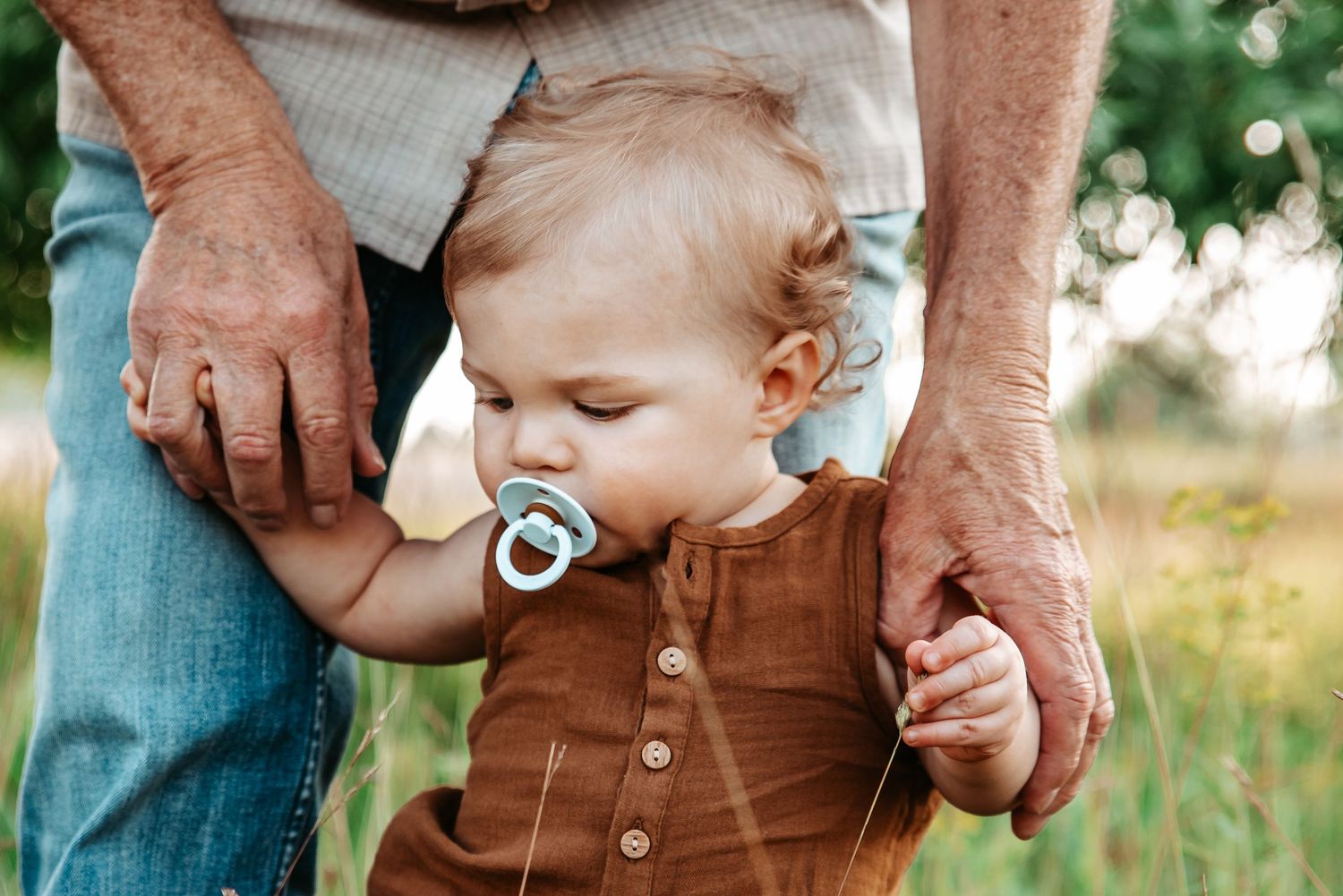 Toddler in brown overalls holds adult's hands while walking outdoors.