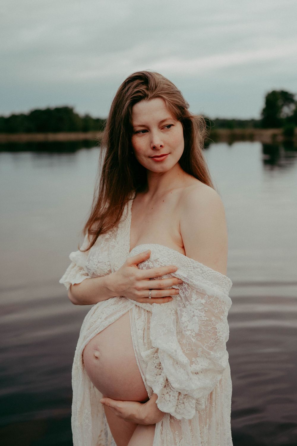 Artistic maternity portrait featuring delicate white lace against a moody lakeside backdrop.