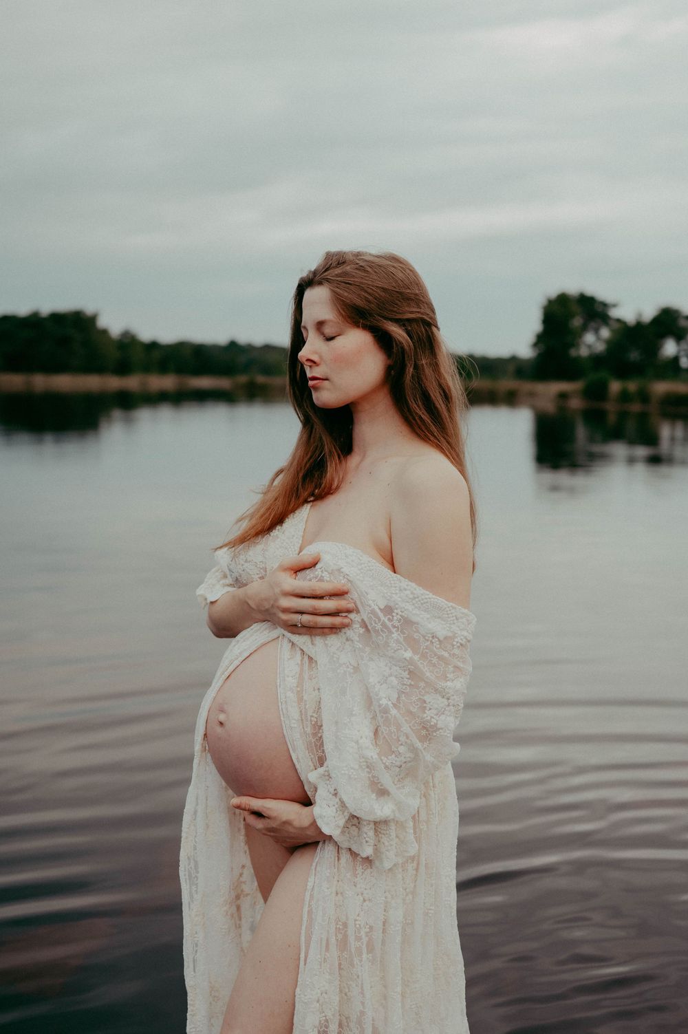 Ethereal maternity photo of an expectant mother in white lace standing in a serene lake during overcast weather.