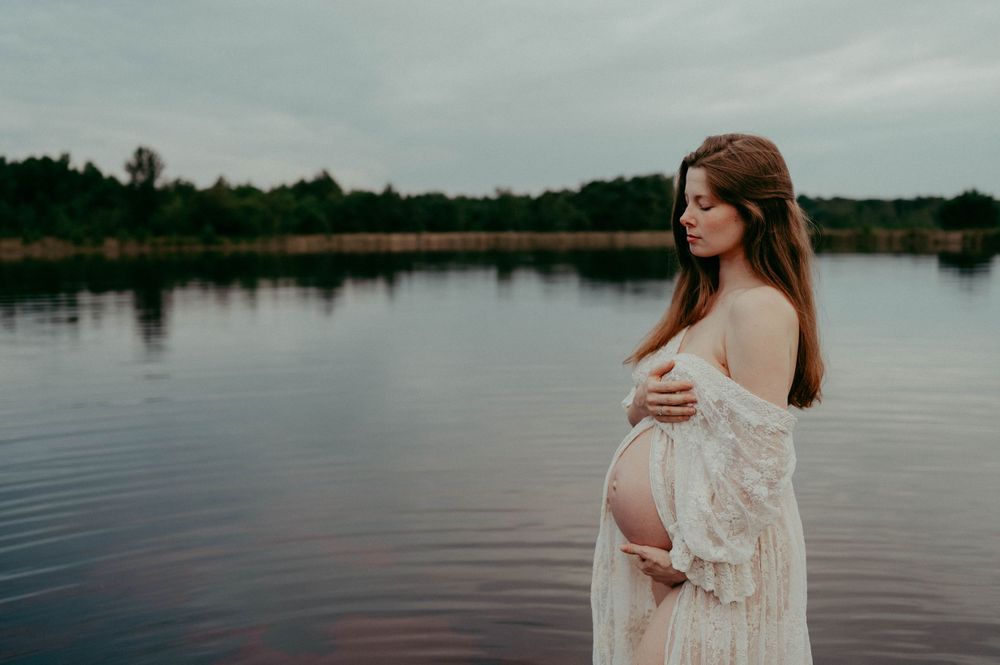 Elegant maternity silhouette against calm waters and overcast sky showing a tender moment.