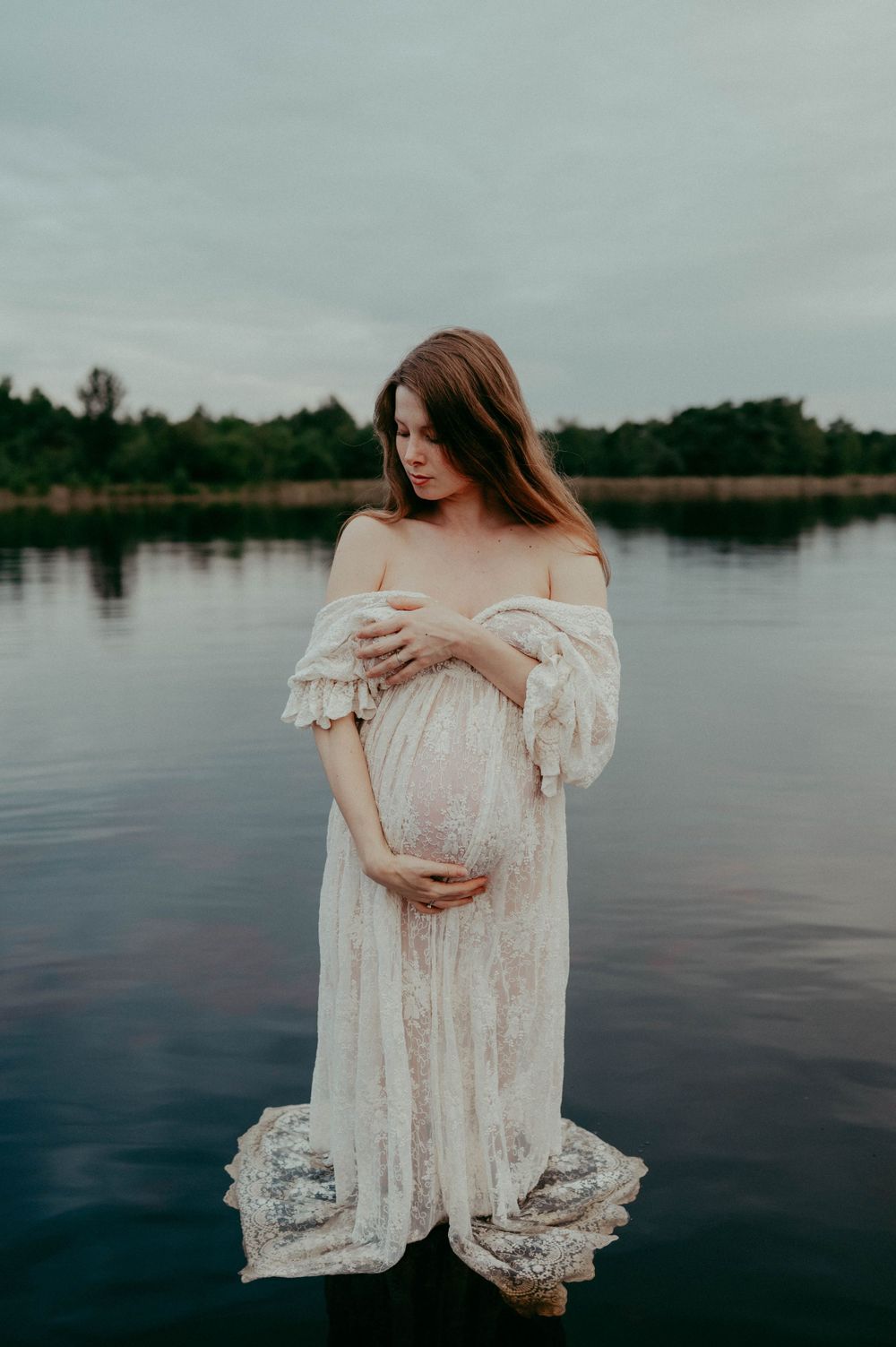 Atmospheric lakeside maternity photo featuring vintage-inspired lace dress and moody sky.