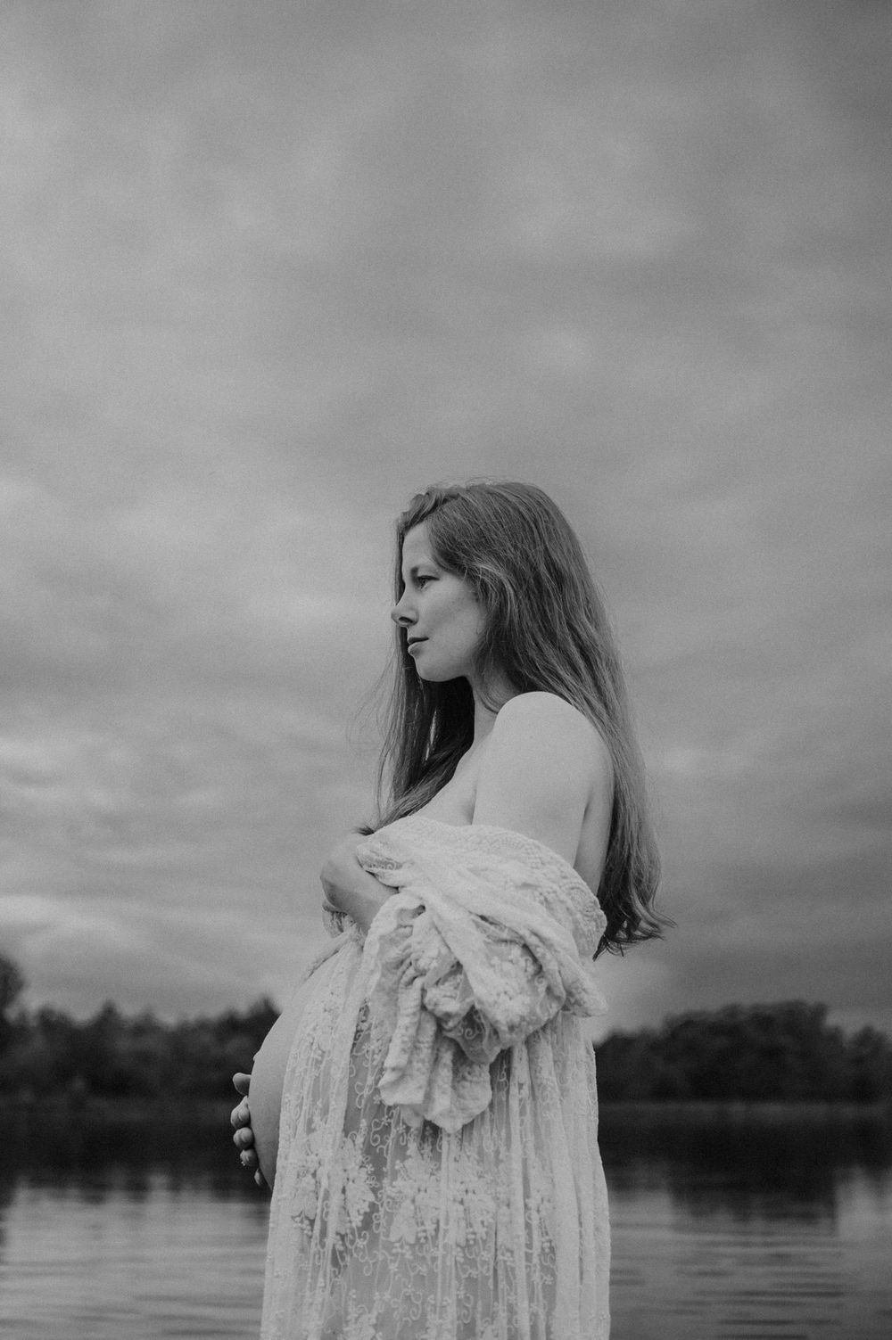 Black and white maternity portrait showcasing flowing lace dress against dramatic cloudy sky.