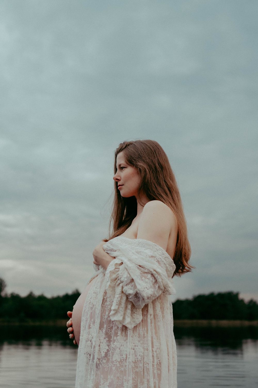 Moody maternity portrait with delicate lace details against peaceful lake scenery.