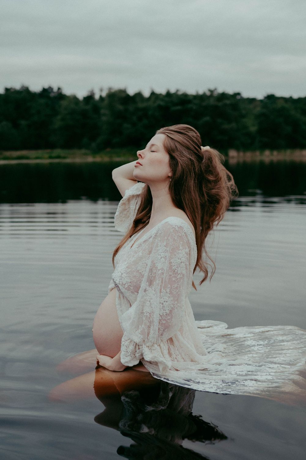 Artistic water maternity photo with flowing lace dress and serene lake backdrop.