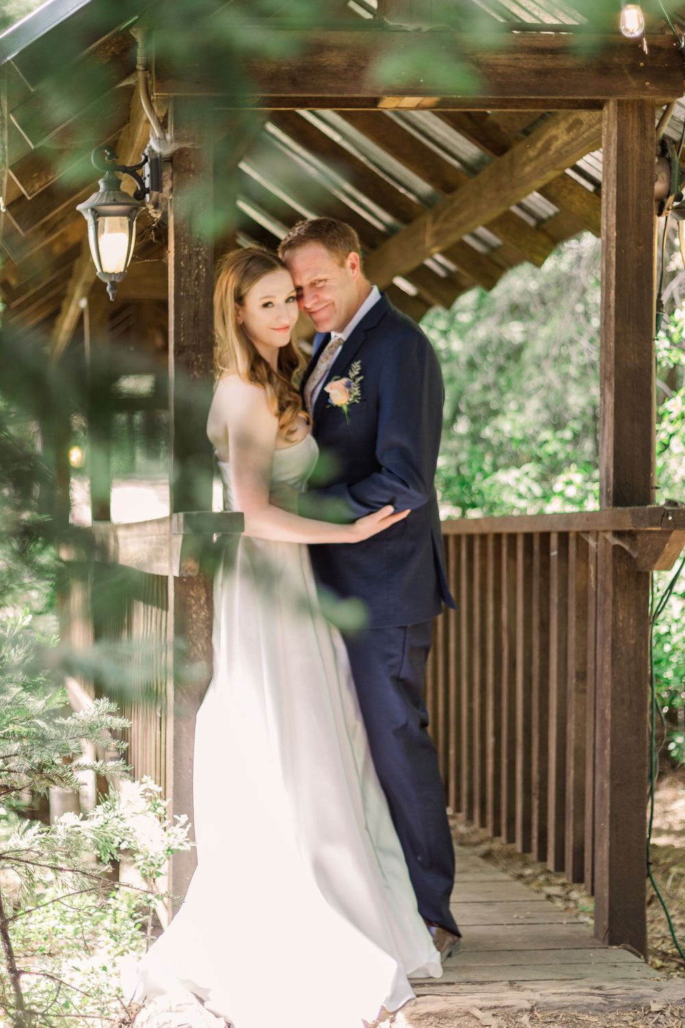 Romantic wedding couple embracing under a rustic wooden covered bridge in a forest setting.
