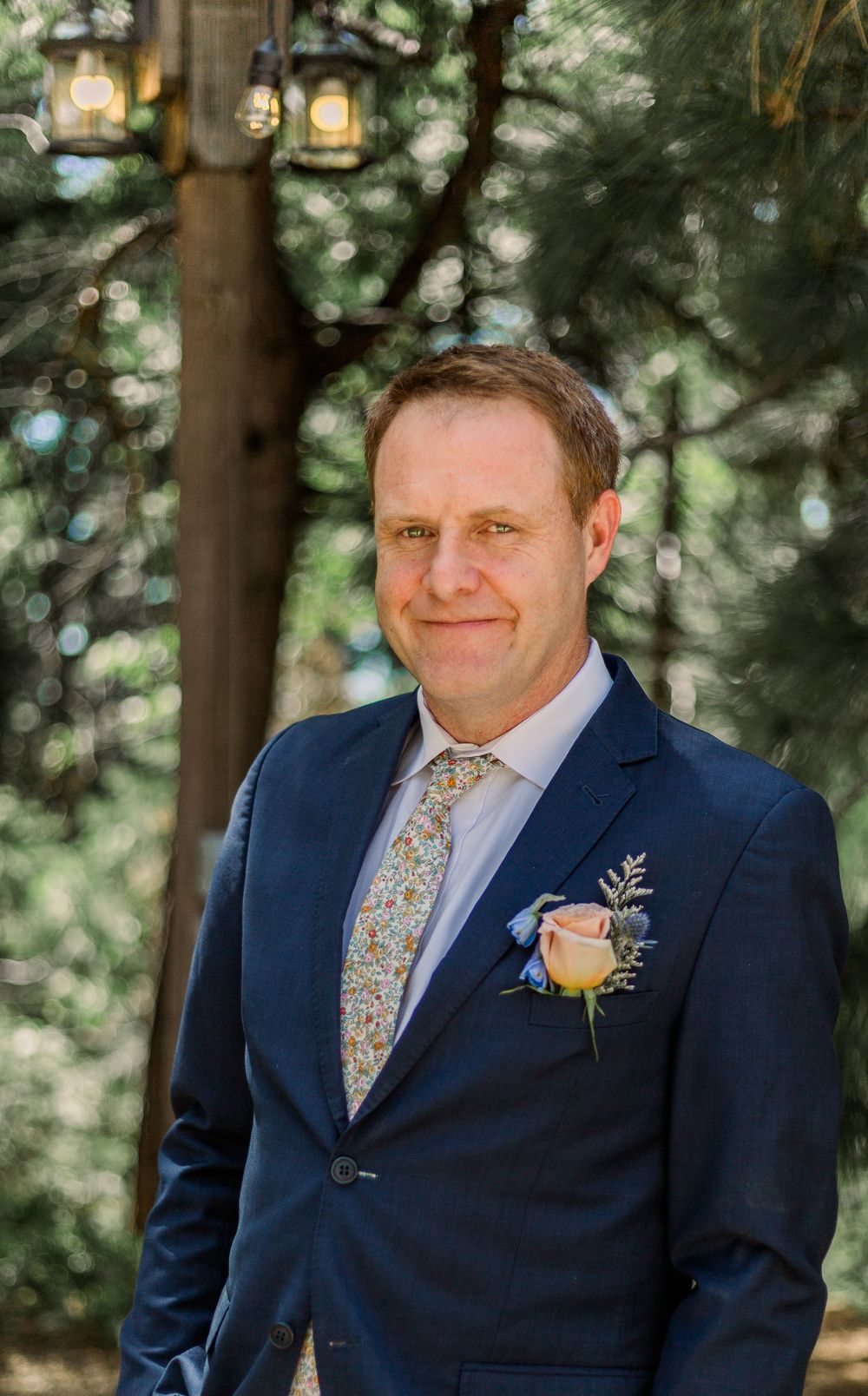 A formal portrait in a navy suit with floral boutonniere against a forest backdrop.