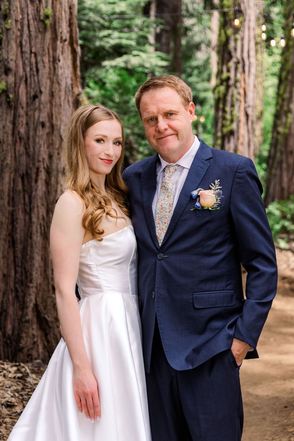 A couple poses for romantic wedding portraits on a forest path surrounded by towering trees.