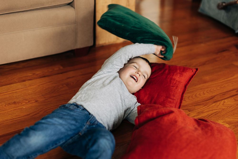 A child lies on hardwood floor surrounded by colorful pillows.