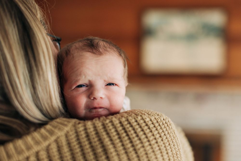 A baby rests against a brown knit sweater in warm indoor lighting.