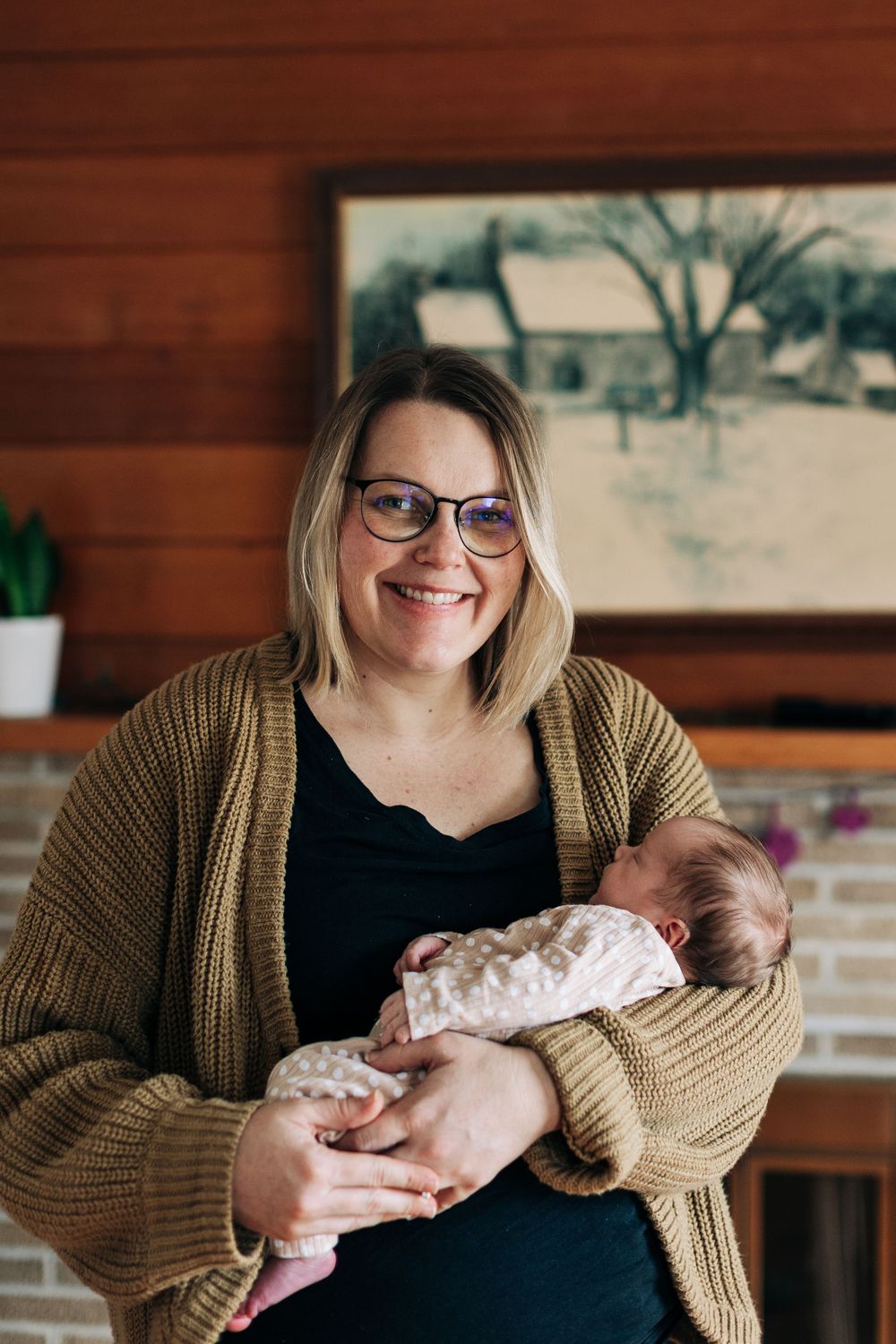 Joyful indoor portrait while cradling a sleeping newborn baby wrapped in white.