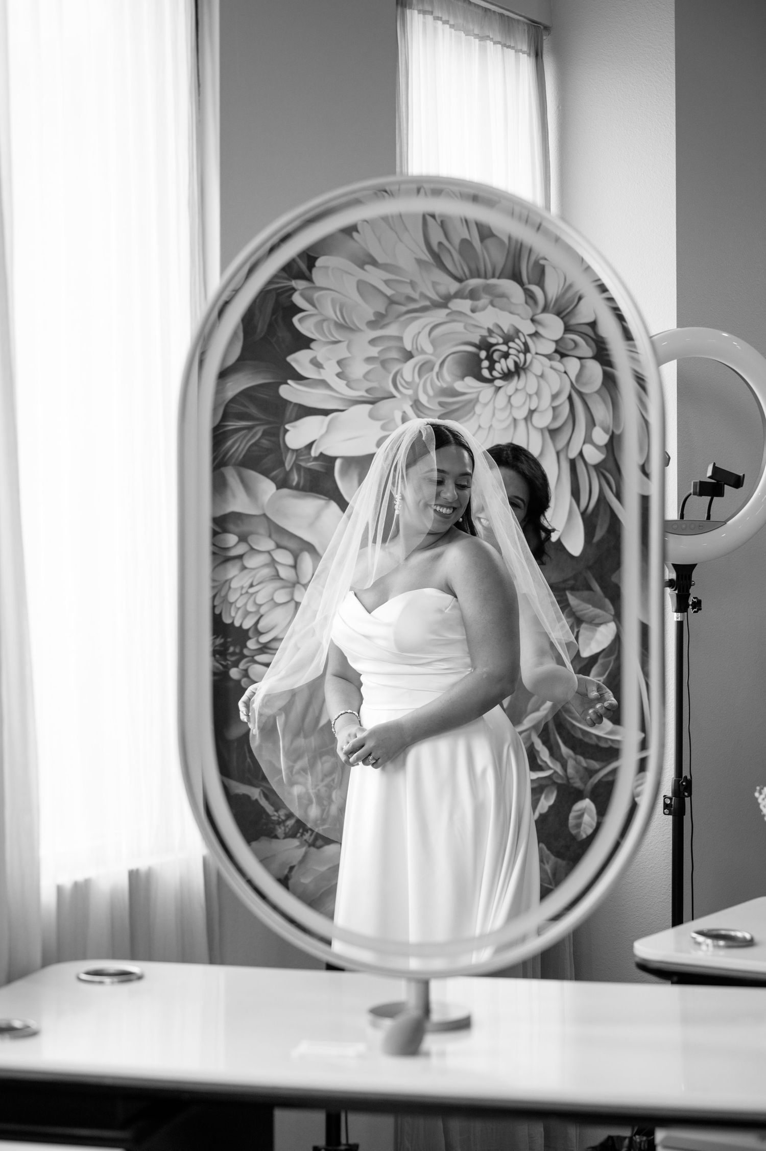 A wedding dress reflection in an oval mirror with a large floral mural backdrop in black and white photography.