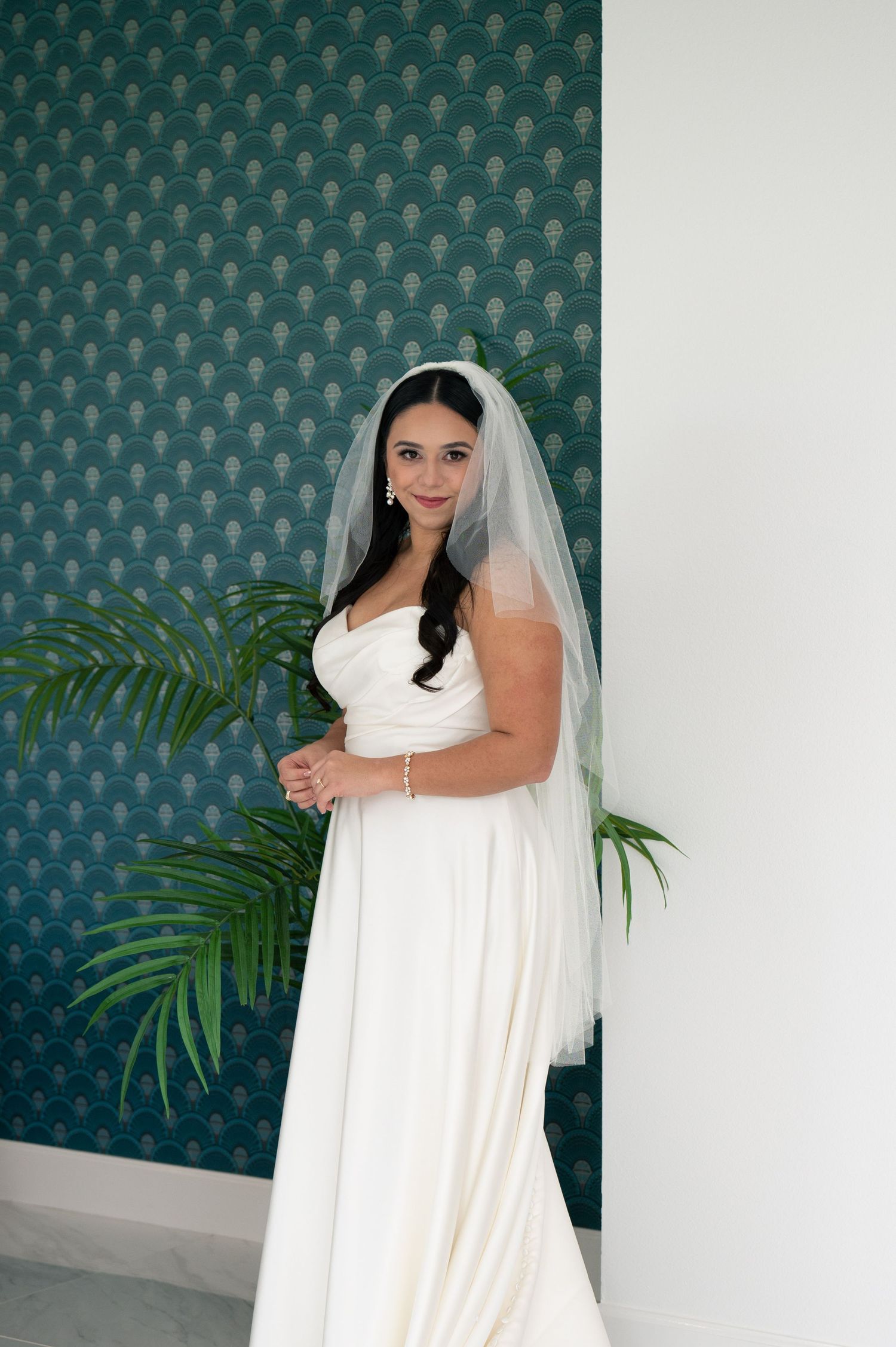 A bride in an elegant white wedding gown and veil poses against a teal floral wallpaper with palm fronds.