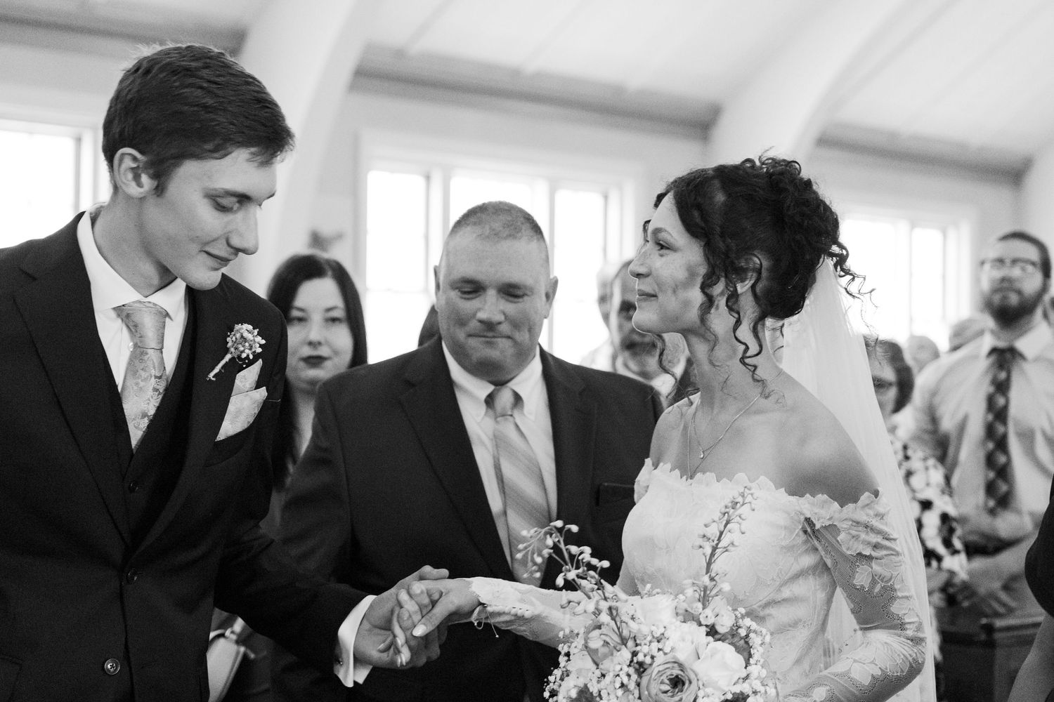 Black and white wedding photo of groom placing ring on brides finger at altar with officiant and guests.