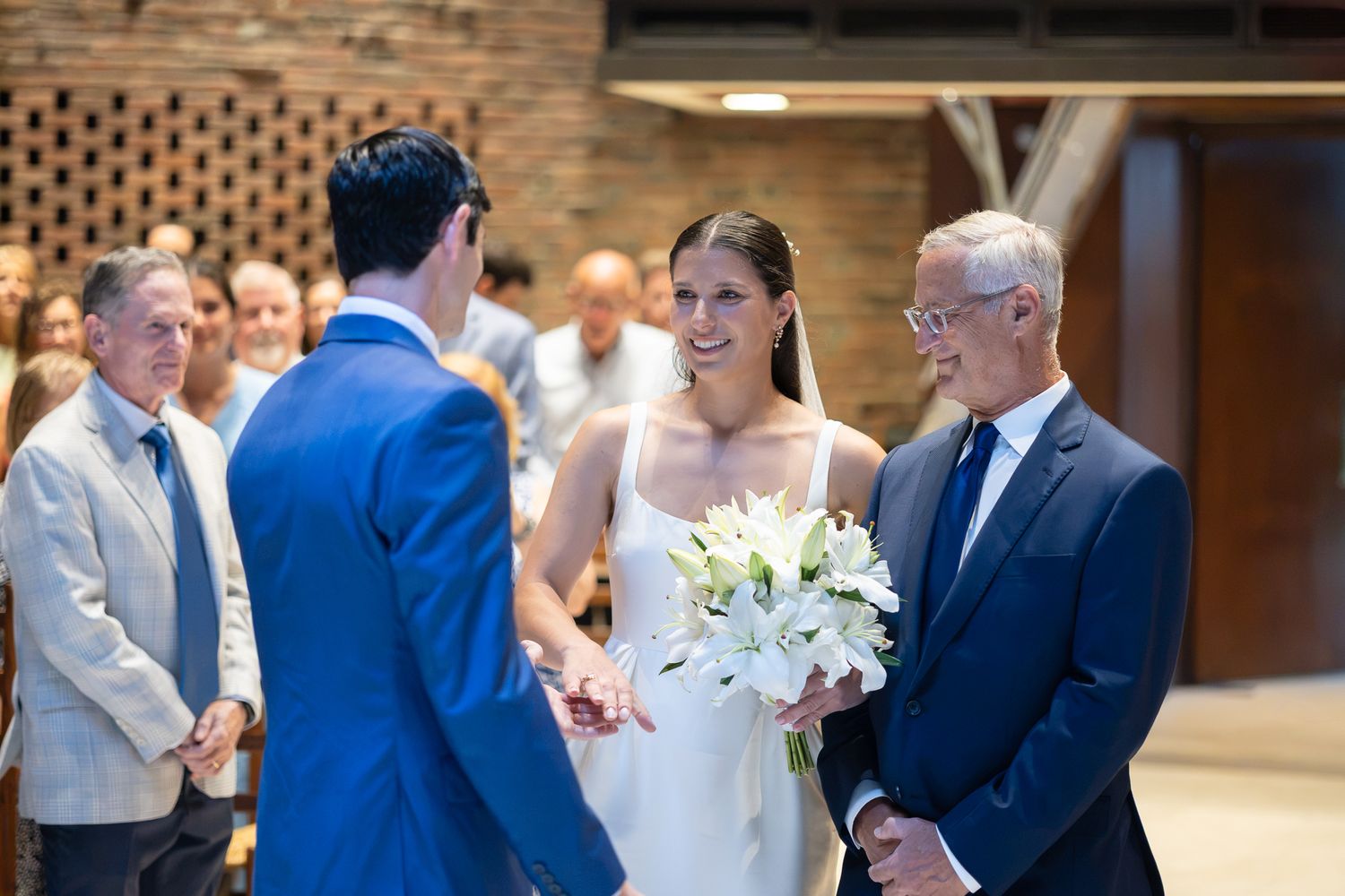 Bride holding white lily bouquet smiles at groom during indoor wedding ceremony with guests seated in background.
