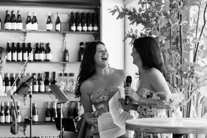 Two women in elegant dresses laughing and sharing a microphone at a wine bar celebration.