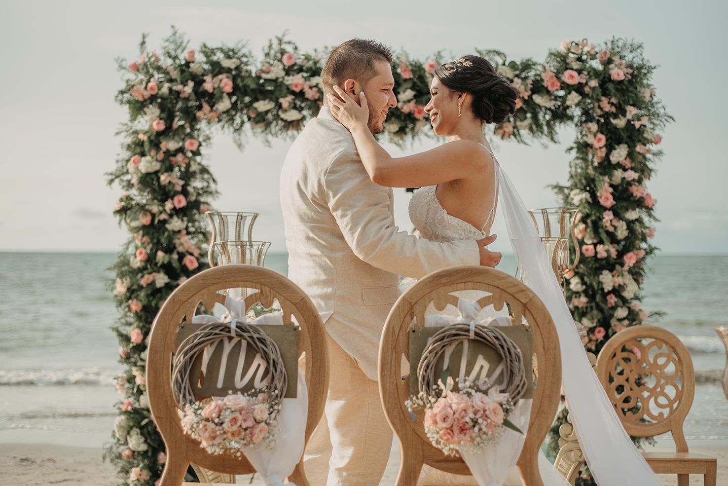 Bride and groom share romantic moment under floral arch adorned with pink roses on beach.