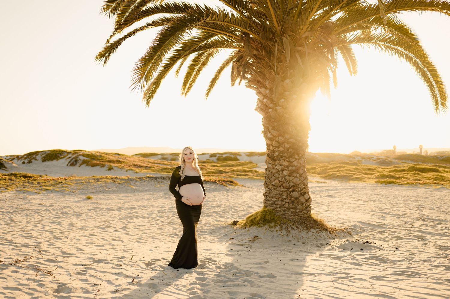 pregnant woman in black two piece dress exposing her bare belly, posed on Oxnard Shores beach for her Ventura maternity photoshoot