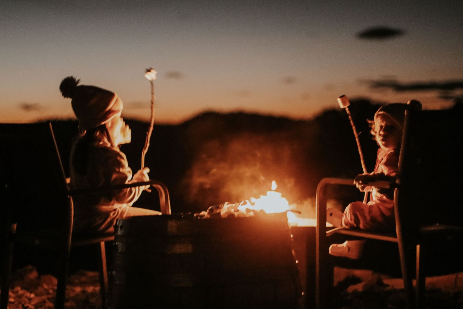 Two children roasting marshmallows over a glowing campfire at dusk, wearing cozy winter hats.